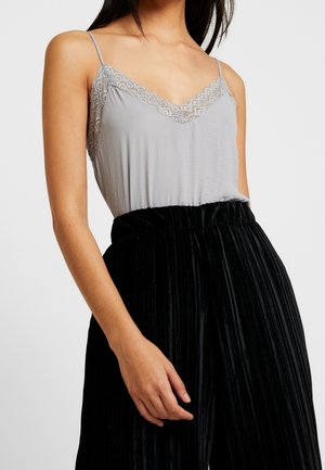 Camisole grise avec décolleté en dentelle et fines bretelles, associée à un pantalon noir plissé taille haute. Les textures des tissus contrastent subtilement.