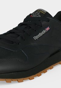 Zapatilla Reebok Classic de piel negra con cordones negros, logotipo de la bandera Union Jack y suela de goma.