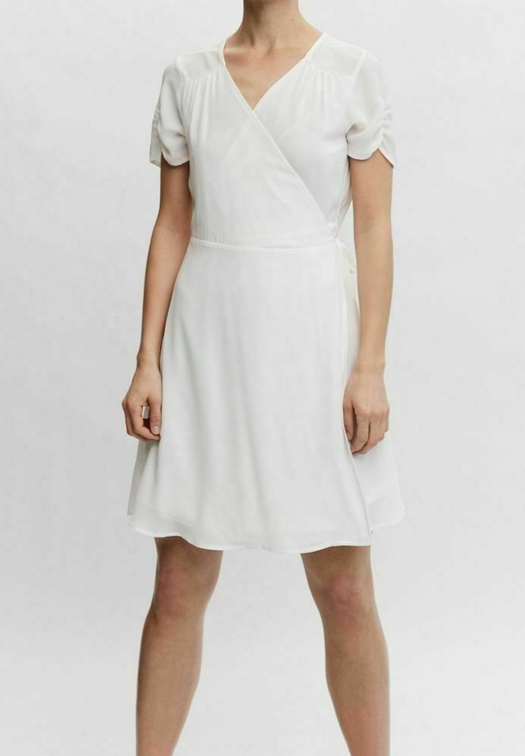 Femme portant une robe portefeuille blanche à manches courtes avec manches froncées et un ourlet à hauteur des genoux, debout devant un fond clair uni.
