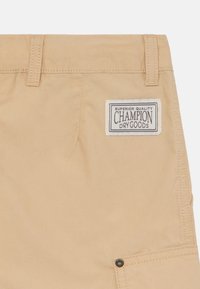 Des shorts beige en tissu léger et lisse avec une taille cousue, un patch logo portant la mention "Champion Dry Goods", et un accent en bouton en métal.
