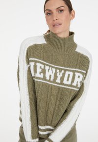 Olivgrüner Strickpullover mit hohem Kragen, weißen Zopfstrickärmeln und "NEW YORK"-Aufschrift auf der Brust in Weiß.