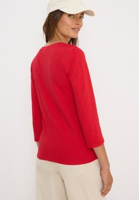 Rotes Langarmoberteil mit rundem Halsausschnitt und Dreiviertelärmeln, aus einem glatten Baumwollgemisch. Getragen mit beigen Hosen.