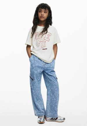 Light beige t-shirt with red lettering, paired with light blue denim pants featuring a subtle pattern. Model wears silver shoes.