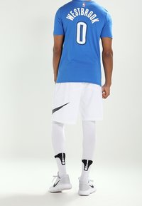 Niebieski trykot z napisem „WESTBROOK” i numerem „0” na plecach, w połączeniu z białymi szortami, białymi legginsami kompresyjnymi i szarymi sneakersami marki Nike.