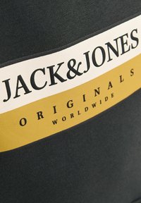 Tela negra con una textura suave que presenta una etiqueta blanca y amarilla. La etiqueta tiene "JACK&JONES" en texto negro en negrita y "ORIGINALS WORLDWIDE" abajo.