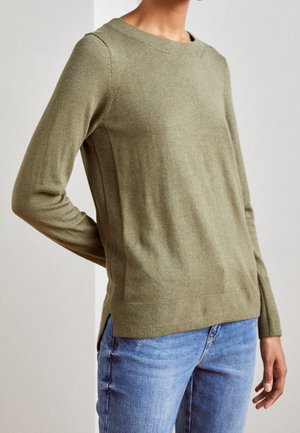 Olivgrüner Strickpullover mit Rundhalsausschnitt, langen Ärmeln und seitlichen Schlitzen. Weiche Textur mit glatter Oberfläche. Kombiniert mit blauen Jeans.