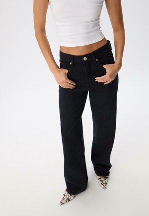 Femme portant un jean noir taille haute à jambes larges, un top court blanc, les mains dans les poches, et des talons aiguille à imprimé animal sur un fond blanc.