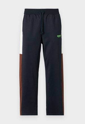 Marineblauwe trainingsbroek met witte en bruine zijpanelen, elastische tailleband, trekkoord en een groene logo op de heup. Zacht, glad materiaal.