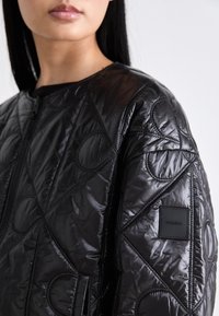 Vrouw in een zwarte gewatteerde jas met geometrische en cirkelvormige stitching en een Calvin Klein logo patch op de mouw.