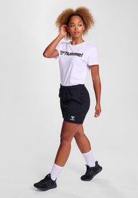 Hvit t-skjorte med svart "Hummel" logo, kombinert med svarte shorts. Modellen har på seg svarte joggesko og hvite sokker. Enkel, avslappet design.
