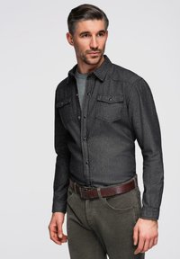 Chemise boutonnée gris foncé avec deux poches poitrine, un col, et une coupe ajustée, associée à un pantalon vert olive et une ceinture en cuir marron.