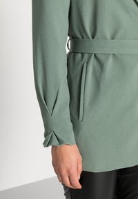 Blusa verde con maniche lunghe, una cintura legata in vita e tessuto texture. Dettaglio arricciato ai polsini delle maniche. Liscia e leggera.