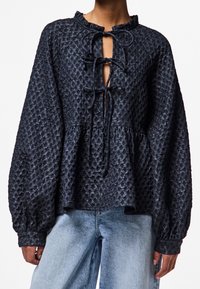 Blouse texturée bleu marine avec un col à volants, ouverture frontale avec liens, manches longues bouffantes et ourlet à basque, confectionnée dans un tissu léger.