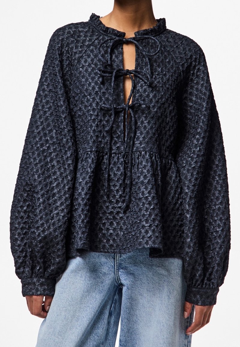 Blouse texturée bleu marine avec un col à volants, ouverture frontale avec liens, manches longues bouffantes et ourlet à basque, confectionnée dans un tissu léger.