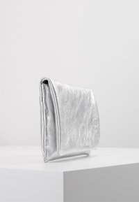 Pochette argentée brillante posée debout sur une surface blanche, sur un fond gris clair uni.