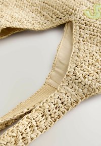 Beige vävd väska gjord av naturfiber med mjukt, slätt tygfoder. Har tjocka remmar och diskret grön broderad detalj.