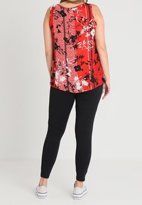 Rotes Blumenshirt ohne Ärmel mit schwarzen Akzenten und weißen Mustern, kombiniert mit schwarzen Leggings und weißen Sneakers. Glattes Material, lässiges Design.
