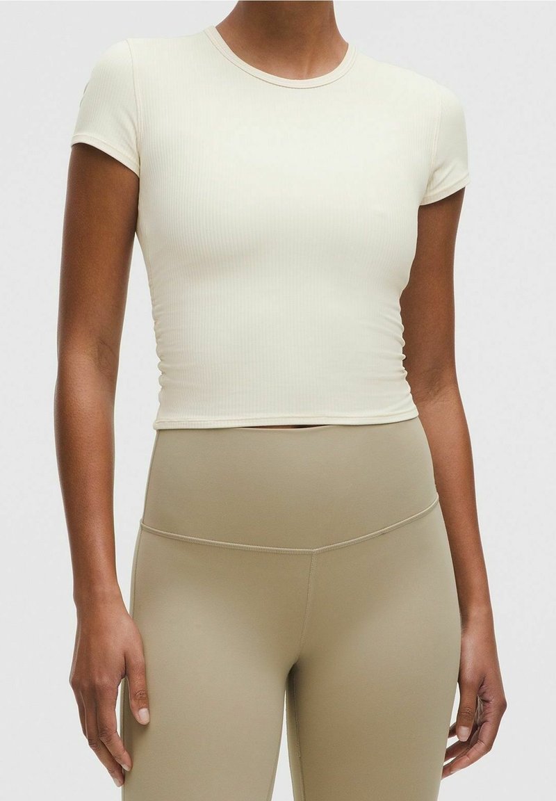 Femme portant un crop top blanc ajusté à manches courtes et un legging beige taille haute sur un fond uni.