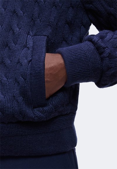 Main à la peau foncée insérée dans la poche d'un pull épais bleu marine en maille torsadée avec poignets et ceinture côtelés.