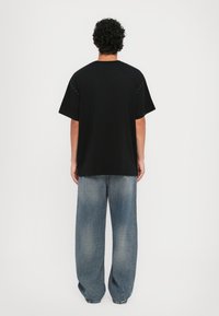 Svart T-skjorte med kort erme og avslappet passform, med små dekorative detaljer på skuldrene, sammen med løse blå denimbukser.