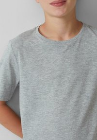 Eine Person trägt ein schlichtes, hellgraues Kurzarm-T-Shirt mit Rundhalsausschnitt und steht vor einem hellen Hintergrund, mit den Armen hinter dem Rücken.