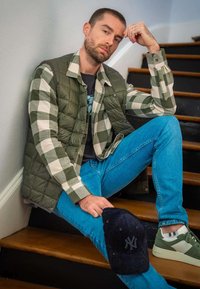 Doudoune sans manches verte olive sur une chemise à carreaux, jeans bleu clair, baskets vertes et une casquette sombre avec un emblème. Assis sur des escaliers.