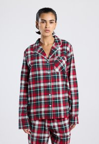 Young person wearing red, green, and white plaid button-up pajama top with matching pants, standing against a plain light background.