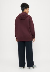 Sweatshirt oversized de cor burgundy com capuz, combinado com calças pretas largas. O conjunto apresenta uma textura confortável e um ajuste descontraído, ideal para uso casual.