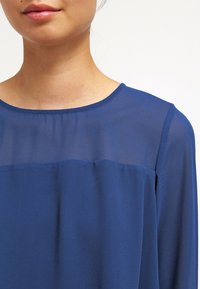 Blå blus med ett semi-transparent överparti och en solid nederdel, rund halsringning och mjukt tyg. Långa ärmar finns.