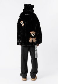 Chaqueta de piel sintética negra con capucha que tiene orejas de oso, adornada con aplicaciones de osito de peluche en dorado y crema, llevada sobre vaqueros oscuros.