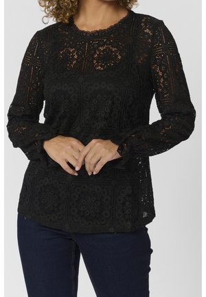 Schwarze langärmelige Spitzenbluse mit filigranen floralen und geometrischen Mustern, getragen zu dunkelblauen Jeans, Hände sanft vor dem Körper verschränkt.