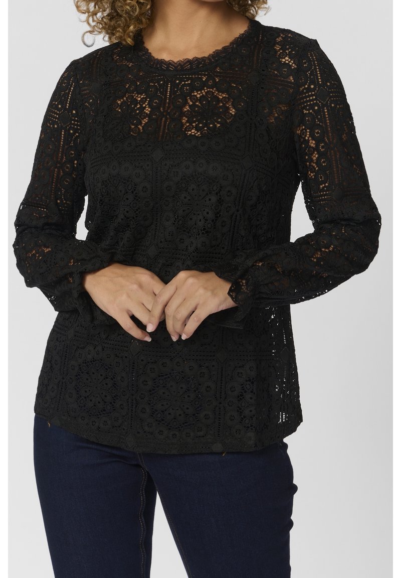 Blouse noire en dentelle à manches longues avec des motifs floraux et géométriques complexes portée avec un jean bleu foncé, mains doucement jointes devant.