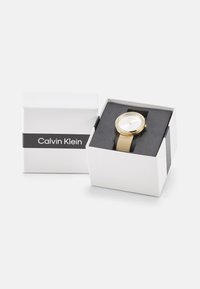 Arany színű óra hálós pánttal, fehér számlappal és kristályos díszítéssel, fehér dobozban, fekete szalaggal és "Calvin Klein" logóval.