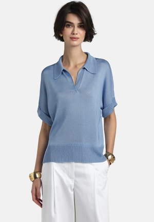 Nő világoskék, rövid ujjú kötött felsőt visel gallérral és fehér, magas derekú nadrágot, arany karkötőkkel és fülbevalókkal kiegészítve.