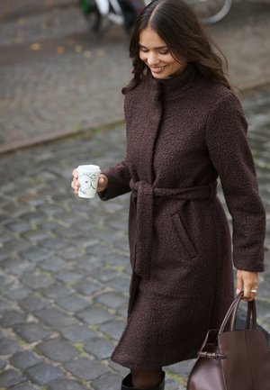 Bruine, gestructureerde jas met een hoge kraag, een ceintuur om de taille en zijzakken. Vrouw die een koffiekopje en een bruine handtas vasthoudt. Achtergrond van kasseien.