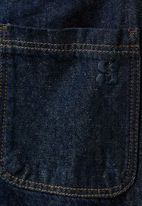 Temno modra jeans tkanina ima zadnji žep s kontrastnim oranžnim šivom in embosiranim logotipom na površini tkanine.