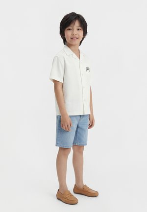 Mladý chlapec stojaci, oblečený v bielej košeli s krátkym rukávom na gombíky, svetlomodrých riflových šortkách a svetlohnedých mokasínach na bielom pozadí.