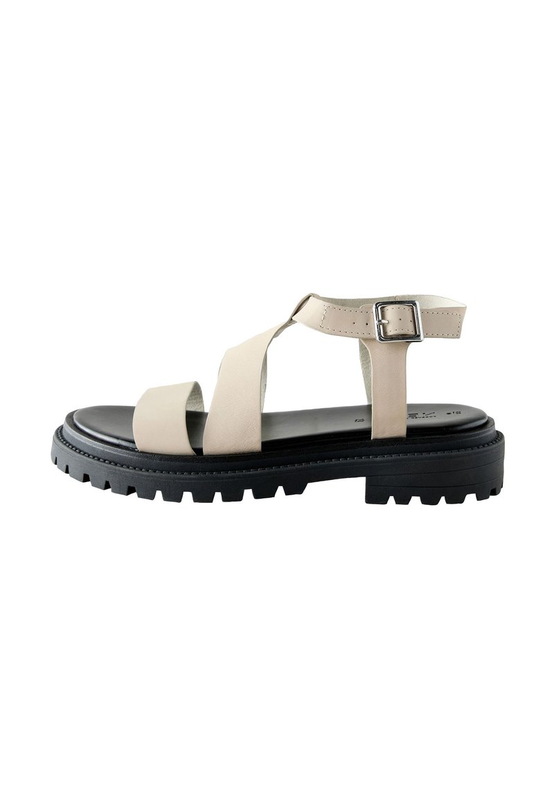Beige lædersandal med justerbar ankelrem og sølvspænde, med en sort kraftig sål med tekstureret slidmønster.