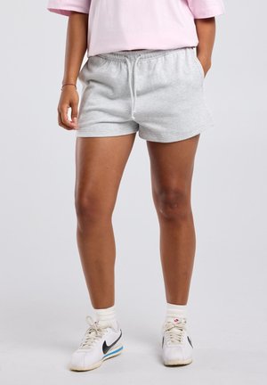 Persoon die lichtgrijze shorts met trekkoord, witte sokken, witte Nike sneakers en een lichtroze T-shirt draagt, staand met de handen in de zakken.