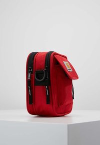 Red fabric bag with two black zippers, a front flap, and a circular ring detail. Features a small logo patch on the front. Compact design.