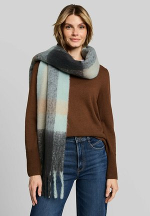 Femme portant un pull marron, un jean bleu et une grande écharpe douce à carreaux gris, bleu et beige enroulée autour de son cou.