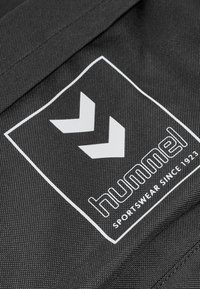 Tela negra con un acabado texturizado que presenta un logo blanco de Hummel, incluyendo un borde rectangular y diseños en forma de flecha.