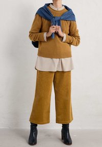 Un pull en velours côtelé moutarde et un pantalon large associés à une chemise beige clair. Un tissu bleu drapé sur les épaules. Des bottines noires.