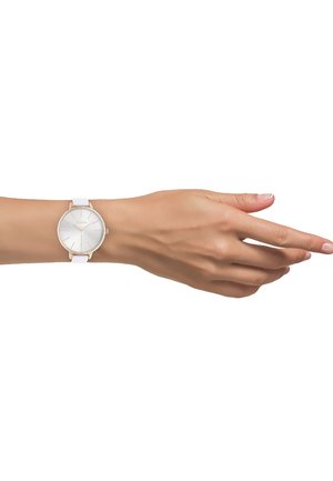 Main féminine à la peau claire portant une montre-bracelet à bracelet blanc avec un cadran blanc et des accents en or rose sur fond blanc.