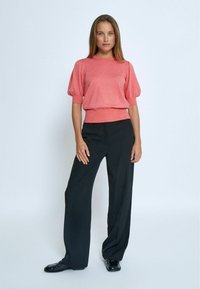 Maglione corto a maniche corte color corallo con collo e polsini a coste, abbinato a pantaloni neri a gamba larga e scarpe nere lucide, modello in piedi su sfondo bianco.