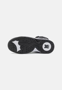 Semelle de chaussure de sport noire avec des motifs de grip texturés, présentant un accent de semelle intermédiaire blanche et un élément de marque circulaire sur le côté.