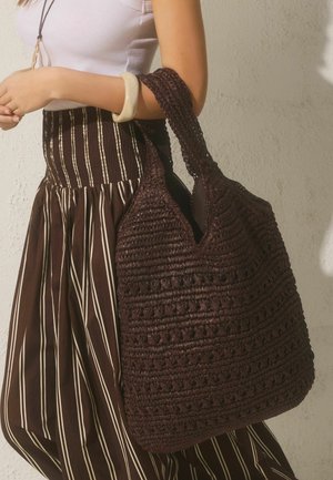 Woman wearing a white sleeveless top and striped brown skirt carries a large dark brown crocheted tote bag on her shoulder.