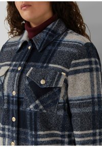 Frau trägt eine blau-grau karierte Wolljacke mit Knöpfen über einem burgunderroten Rollkragenpullover, zeigt Kragen- und Brustdetails.