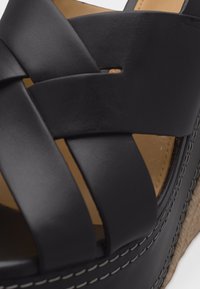 Sandale à semelle compensée en cuir noir avec larges brides croisées et semelle intérieure couleur cuir clair, couture détaillée le long du bord de la semelle.