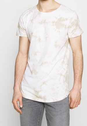 Uomo che indossa una T-shirt a maniche corte tie-dye bianca e beige abbinata a pantaloni grigi, in piedi contro uno sfondo semplice.